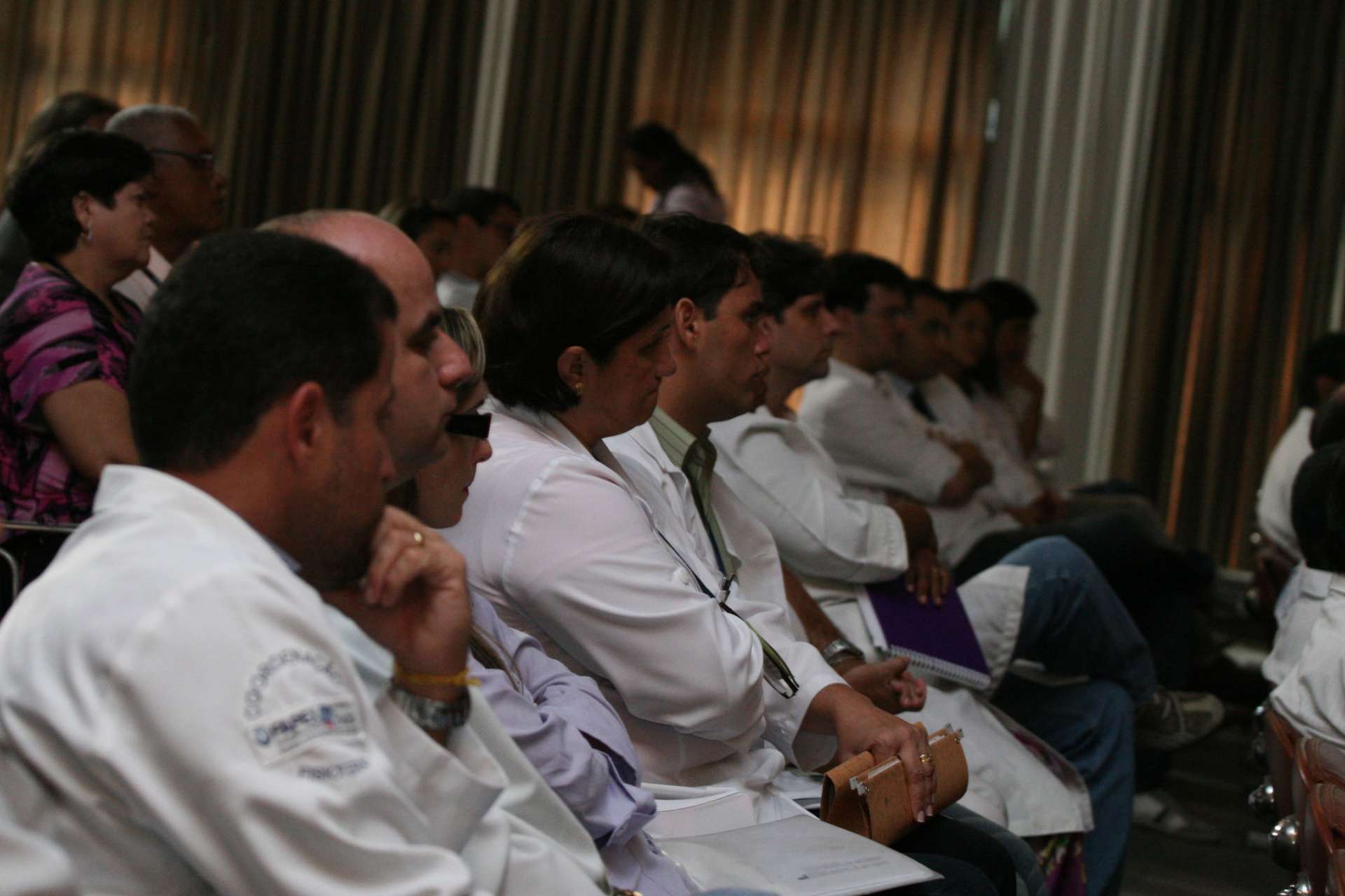 Professionals, many in white coats, listen intently to a Cyber Risk Advisory session in a subdued, curtained conference space.