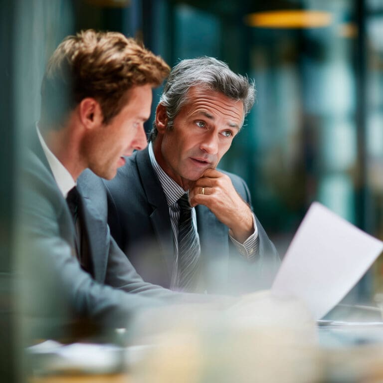 Two executives in suits review a financial services cyber risk document, weighing evidence in an office setting with focused attention.